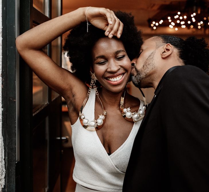 A Black groom kisses a Black bride on the cheek. She wears oversized earrings and a smile on her face.