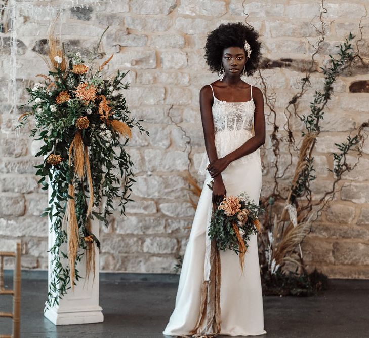 Black bride with afro hair in a lace peplum wedding dress standing by a pillar with orange and green flowers at Ponden Mill