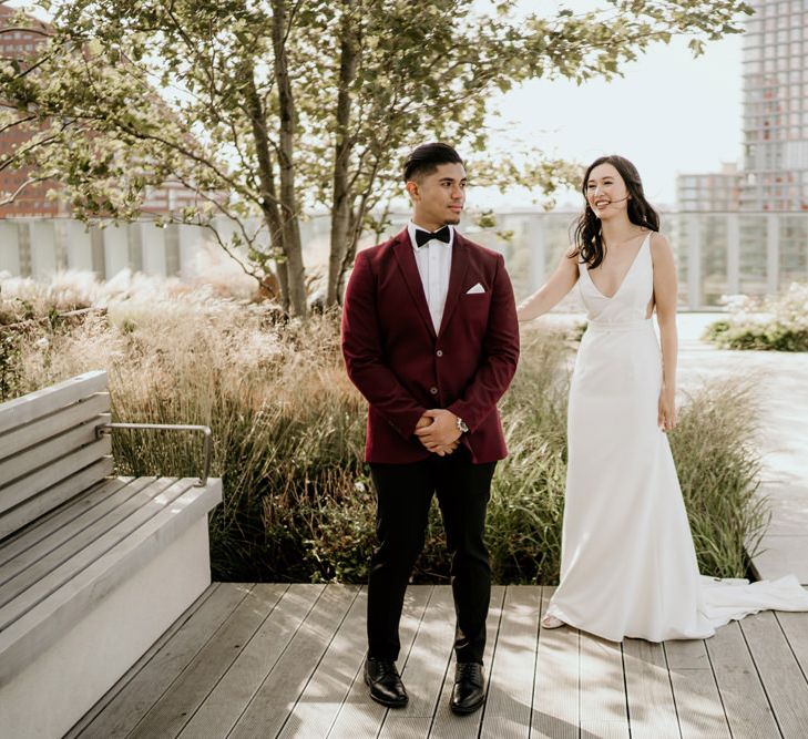 Bride in an A-line wedding dress with straps and plunging neckline approaching her groom in a burgundy tuxedo jacket for their first look