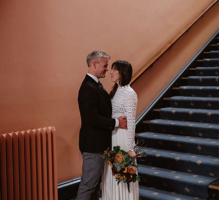 Bride in long sleeved lace top Self Portrait wedding dress holding colourful bouquet stands with groom in brown woollen blazer and waistcoat at Bristol Registry Office