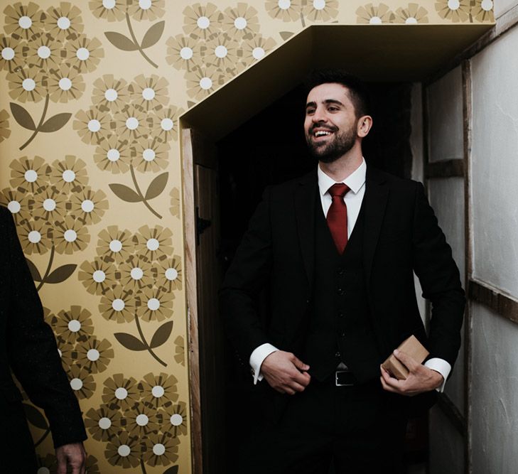 A groom stands in a doorway. He wears a dark suit with red tie.