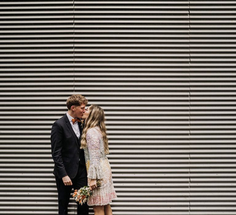 Bride and groom kissing outside steel wall, bride wears colorful pastel dress groom wears three piece suit