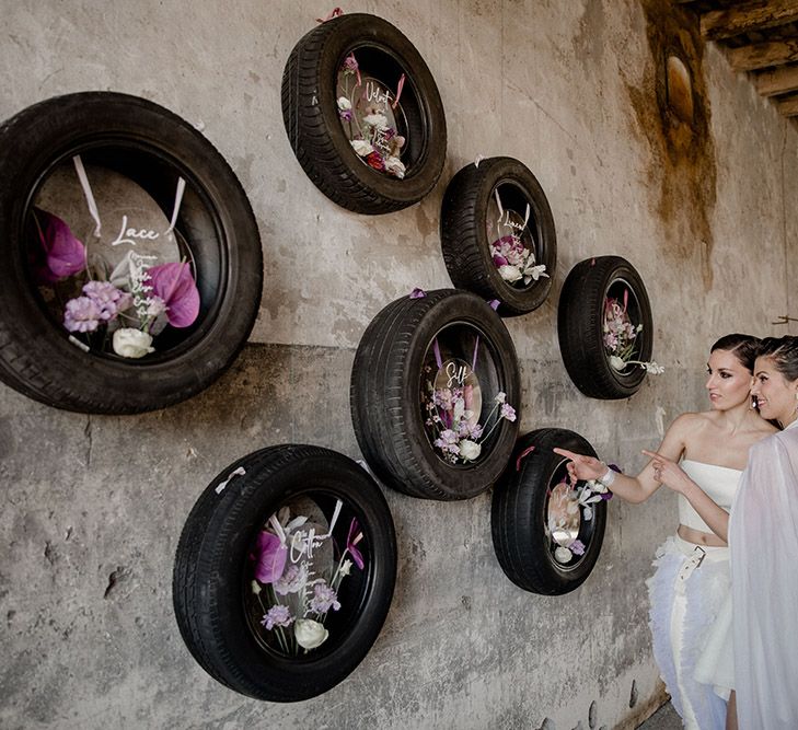 Two brides looking at their tyre table plan at their industrial wedding venue with acrylic signs and purple flowers 