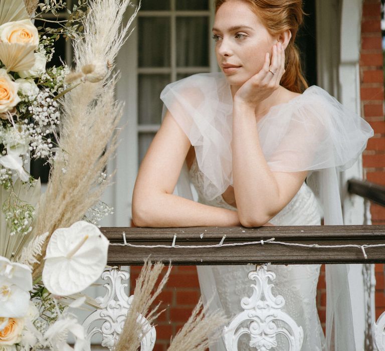 Bride leaning on wooden railing at Berwick Lodge, wearing a puff tulle shoulder detail wedding dress, next to a boho dried flower arch