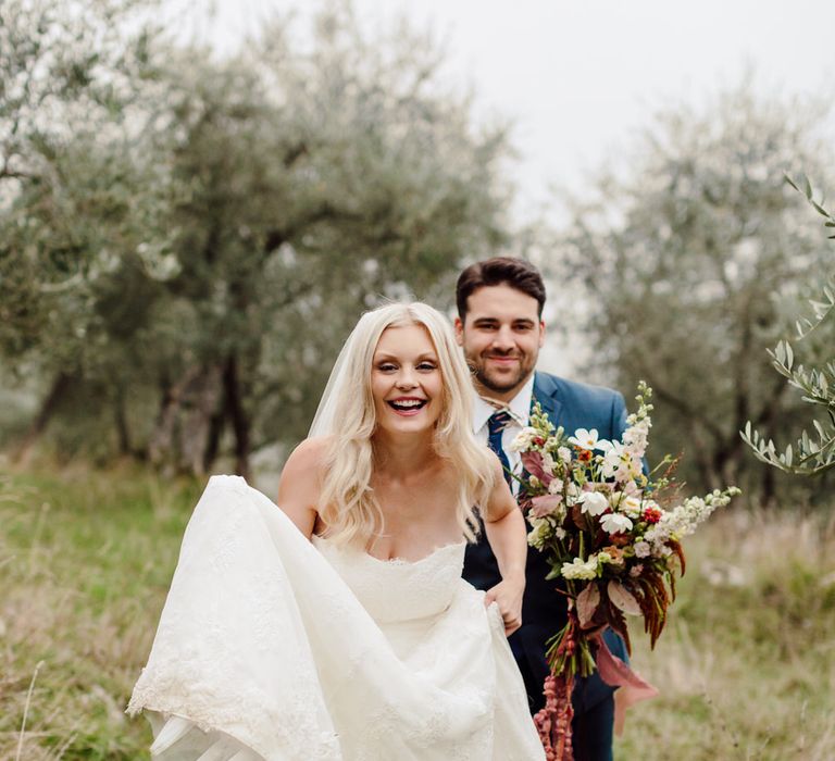 Bride and groom walk through an olive grove in Italy, the groom is holding her bouquet while she lifts up her dress