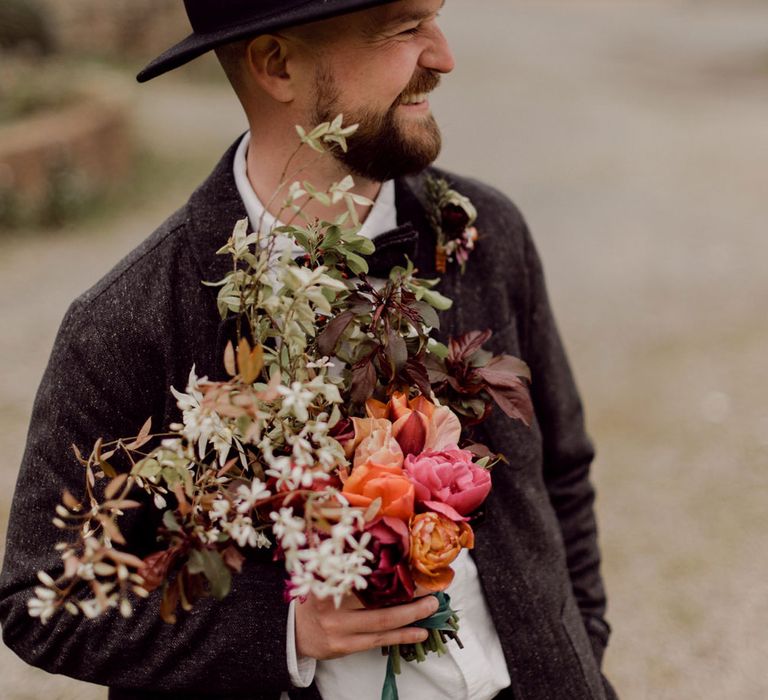 Grom in grey tweed suit and black fedora smiles whilst holding multicoloured wedding bouquet with white flowers and foliage at garden party wedding in Devon