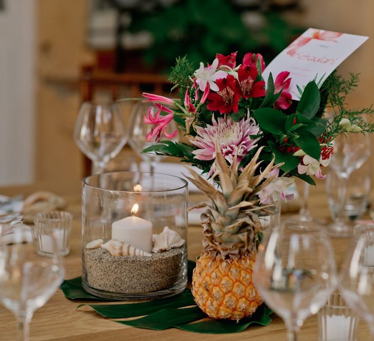 Vibrant pineapple and flower decor with candle in glass holder filled with sand and shells