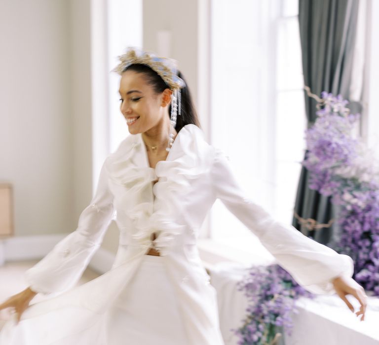 Bride holds out wedding gown with ruffled front whilst stood by lavender trailing from the window