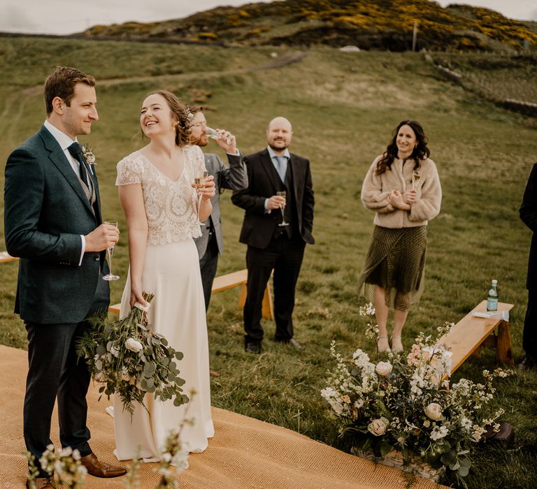 Bride in lace top with capped sleeves and satin skirt smiles at groom in navy suit at clifftop ceremony for Dunluce Castle wedding