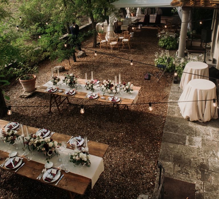 Rustic outdoor reception setting with wooden tables, floral decor and hanging fairy lights within the trees