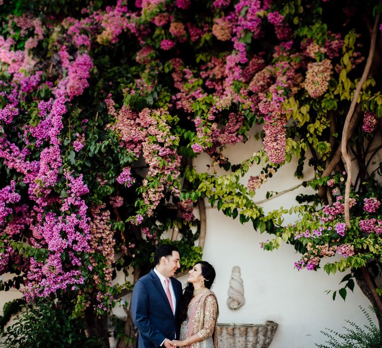 Bride & groom look lovingly at one another as they stand in front of bright pink floral tree as bride wears colourful saree