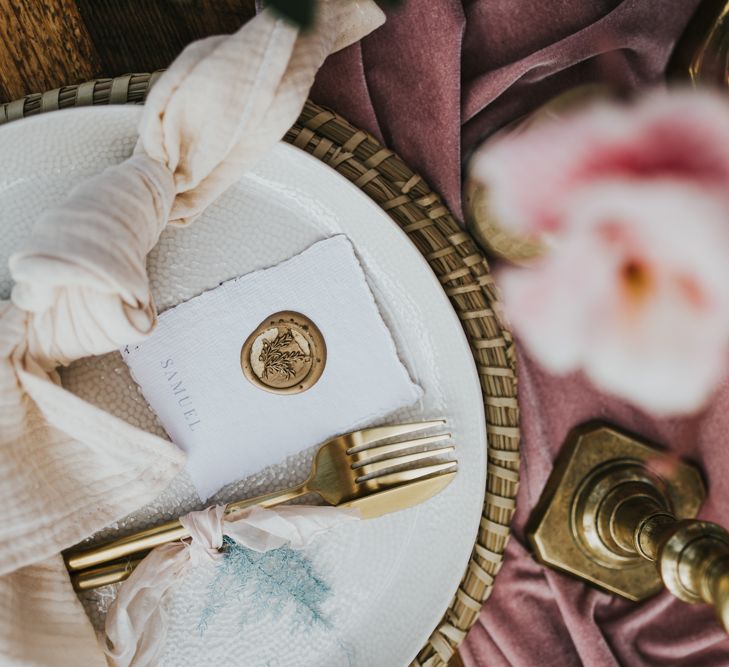 Pastel pink and gold place setting with wicker place mat, gold cutlery, beige linen napkin, and gold wax seal name place card