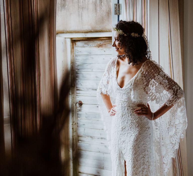 Bride stands with her hands on her hips and wears lace gown with cape as she looks outside door 