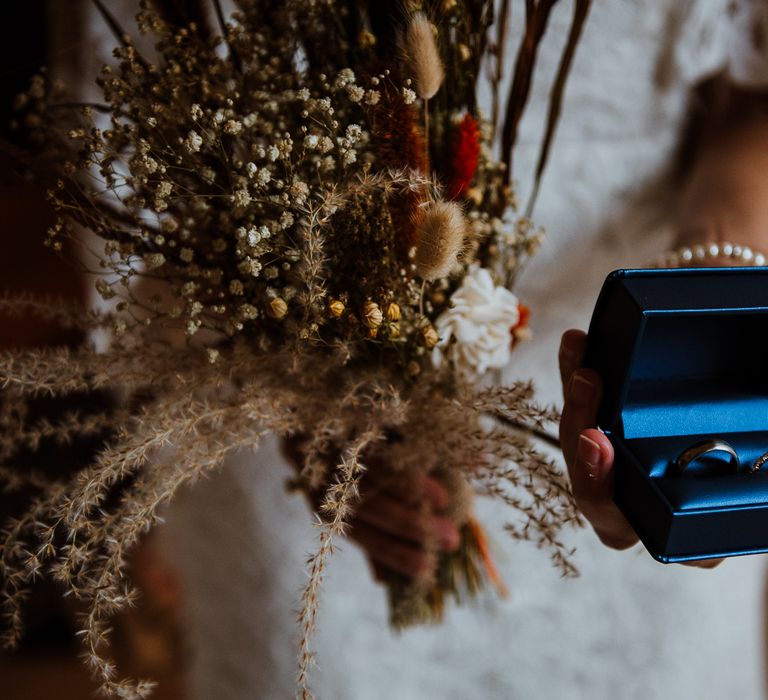 Bride holds out dried floral bouquet and blue box with two silver rings 