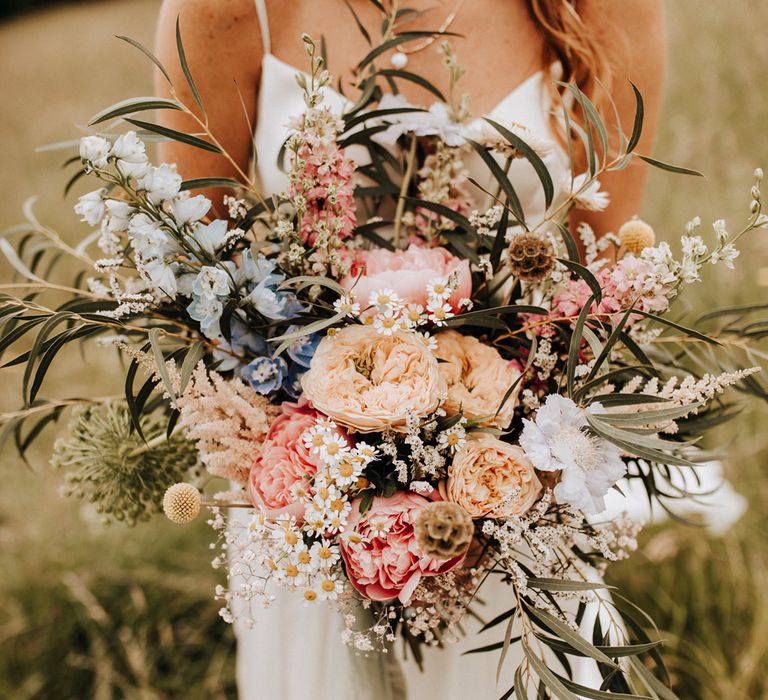 Bride in white cami wedding dress and long curled hair stands in field holding multicoloured bridal bouquet with golden, pink and white flowers