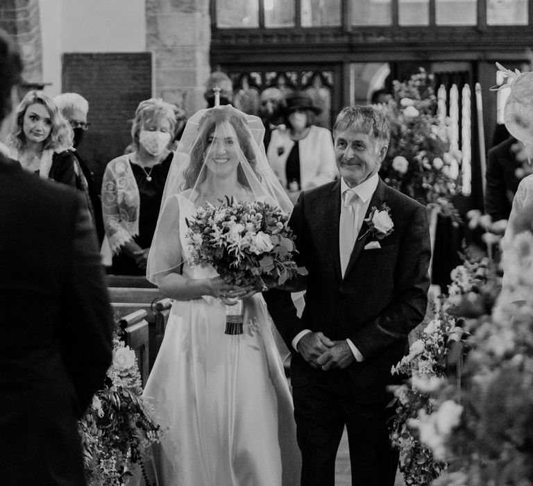 Bride in white Elbeth Gillis gown and veil walks down the aisle arm in arm with father whilst holding white and green bridal bouquet