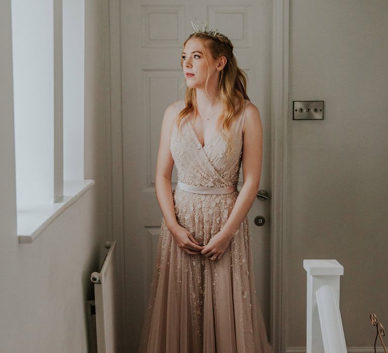 Bride with curled hair and bridal crown stands by window wearing pearl wedding dress before barn wedding cermeony