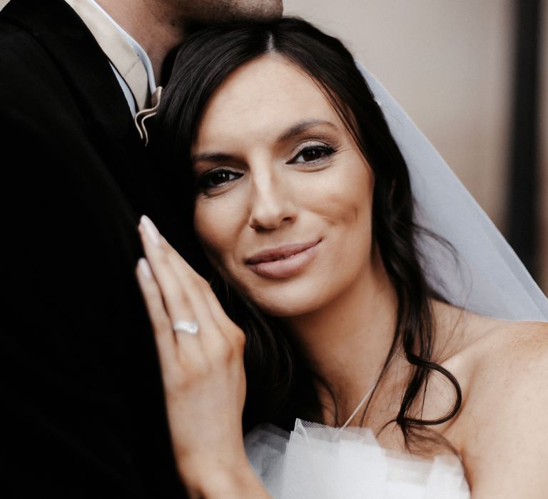 Bride leans into grooms chest as she places her hand on him and looks at the camera whilst slightly smiling