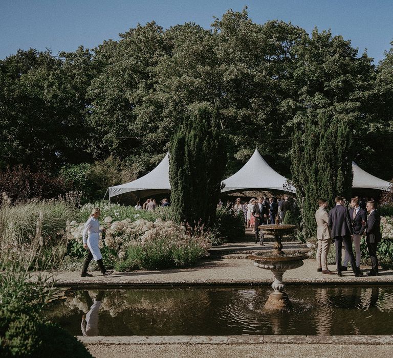 Wedding guests stand in the grounds of Loseley Park by white tent and ornamental pond for summer wedding in Surrey