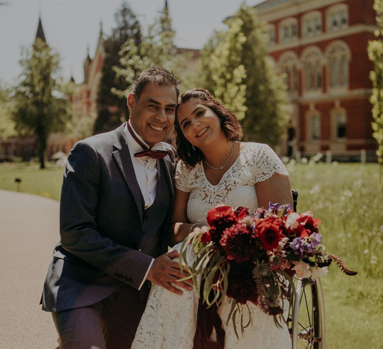 Portrait of an Indian bride and groom at their Dulwich College wedding. The groom wears a blue suit and the bride a lace wedding dress with vibrant bouquet 