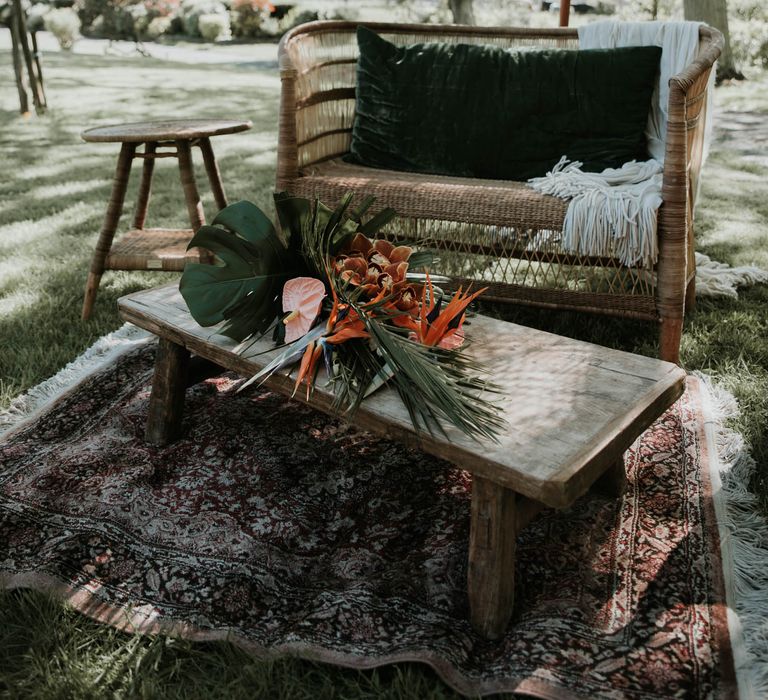 Rattan sofa on vintage rug next to wooden table laid with tropical flowers all under boho tasselled parasol for birds of paradise wedding inspiration