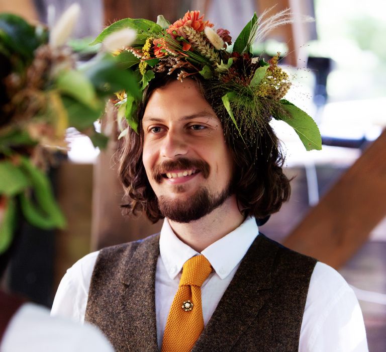 Groom smiles as he wears floral crown and brown waistcoat complete with orange tie