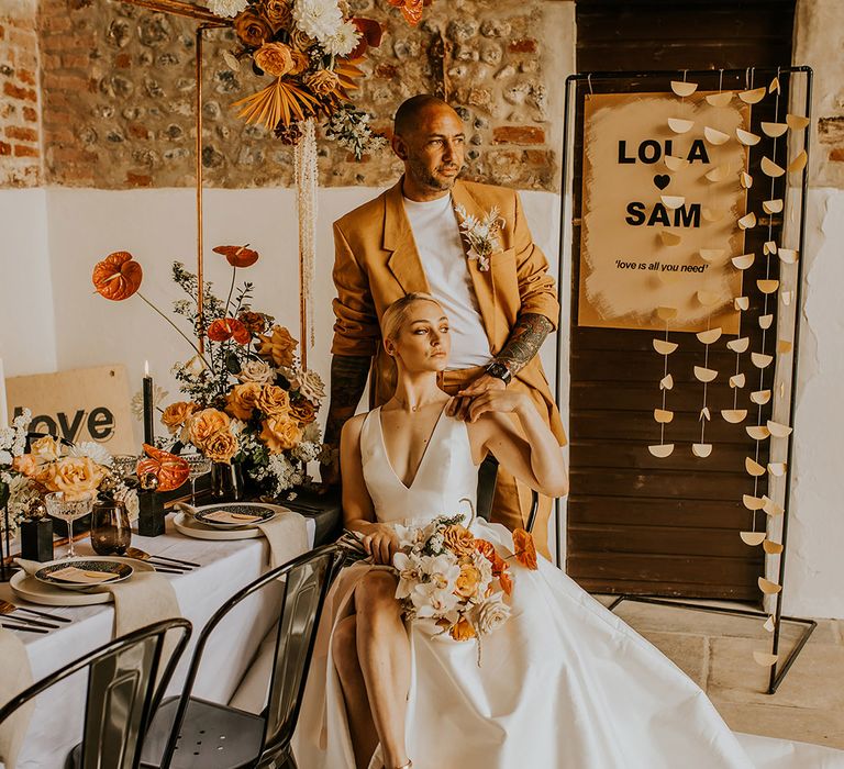 Bride in a Jesus Peiro wedding dress with plunging neckline, front split and long train sitting at her reception table with her groom in a mustard suit by her side 