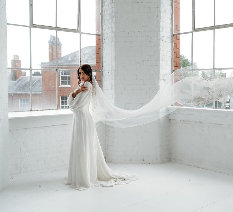 Bride in a floaty ethereal wedding dress with long sleeves wearing a cathedral length embroidered wedding veil in a minimalist industrial wedding venue 