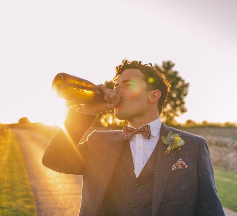 Groom drinks wine from a bottle as the sun sets behind him during golden hour | Story + Colour