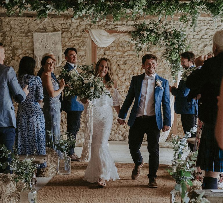 Bride in Grace Loves Lace bardot wedding dress with white and green wedding bouquet walks back down the aisle holding hands with groom in blue suit jacket and floral buttonhole for Isle of Wight wedding with macrame wedding decor