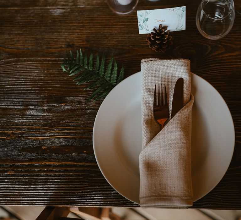 White plate with textured napkins, fern leaf and pinecone table place name on wooden table for rustic tipi wedding reception at Wellington Wood Norfolk