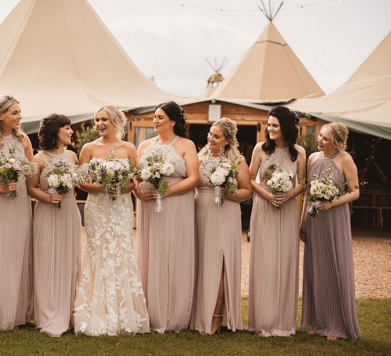 Bride in off the shoulder Enzoani wedding dress stands in the middle of bridesmaids in assorted purple and pink bridesmaid dresses all holding white and green wedding bouquets outside tipi wedding venue at Inkersall Grange Farm