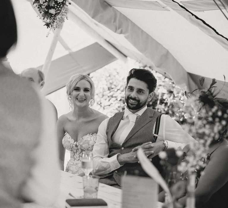 Bride in off the should Enzoani wedding dress and groom in white shirt, white tie and red waistcoat watch guest make speech during tipi wedding reception at Inkersall Grange Farm wedding