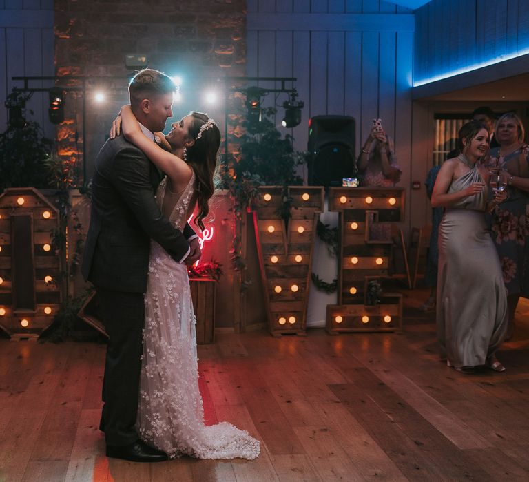 Bride in applique Berta wedding dress and bridal headband dances with arms around groom in grey suit in front of large wooden bulb letters spelling 'LOVE' during wedding reception at Primrose Hill Barn