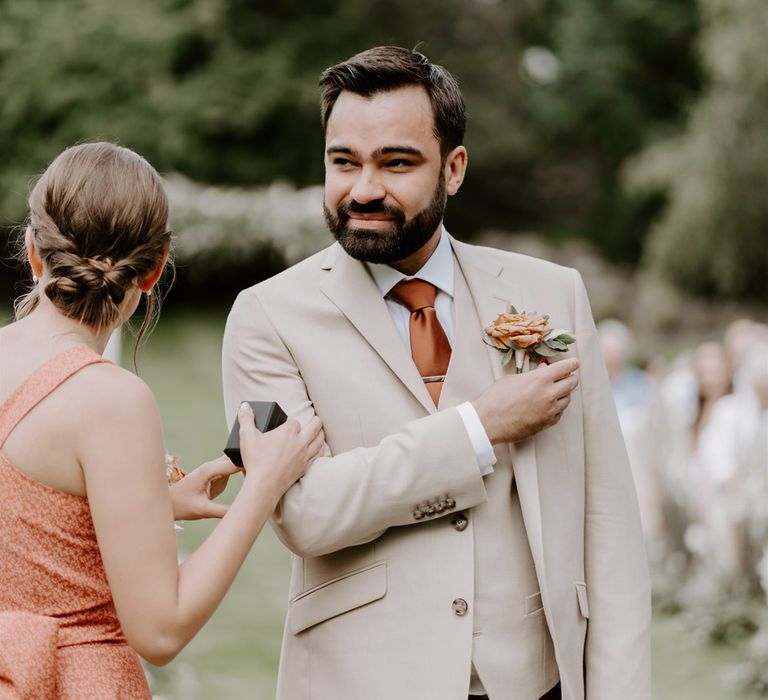 Groom in three piece beige Moss Bros. suit with floral buttonhole waits for bride with bridesmaid in orange dress during DIY garden wedding ceremony in Bedfordshire