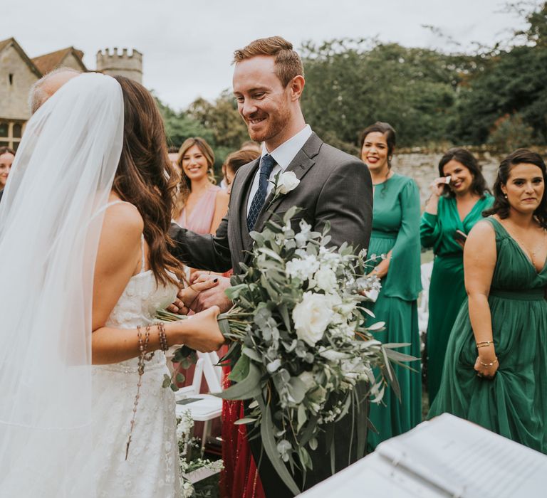 Bride in Pronovias wedding dress and veil holding white rose and green bridal bouquet stands with smiling groom in three piece grey suit during wedding ceremony 