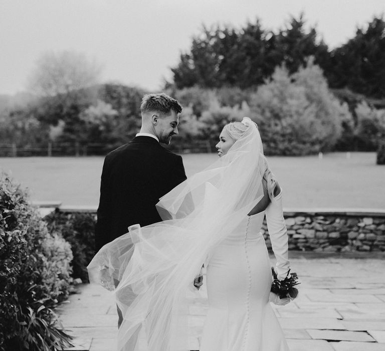 Black and white wedding photograph of the bride in a minimalist fishtail wedding dress with long cathedral length veil blowing behind her 