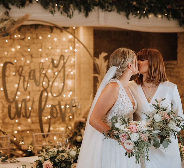 Brides clutching blush bouquets kiss with crazy in love sign backdrop