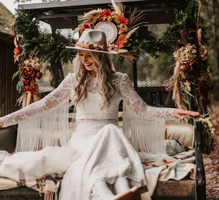 Bride in boho lace tasselled sleeve two piece wedding dress and bridal hat sits outside before wedding