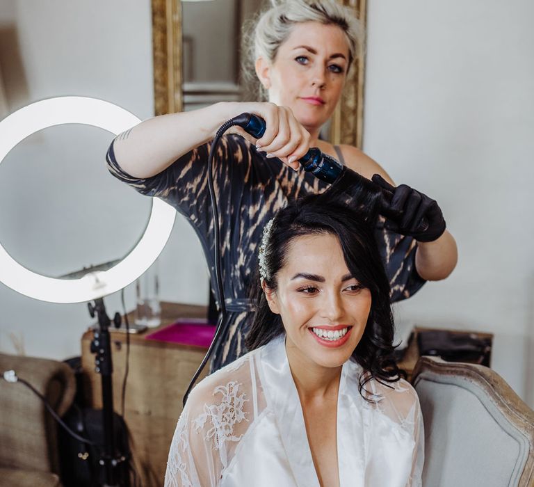 Bride in lace and silk bridal robe holding champagne flute sits having her hair done by hair stylist before wedding