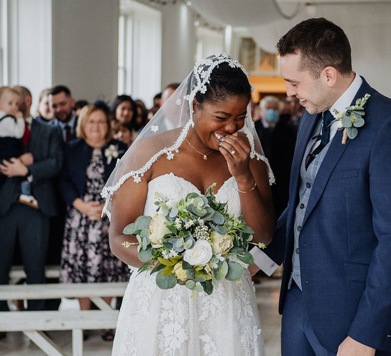 Gorgeous bride smiles with her lace veil with Groom in his fitted navy suit at wedding ceremony 