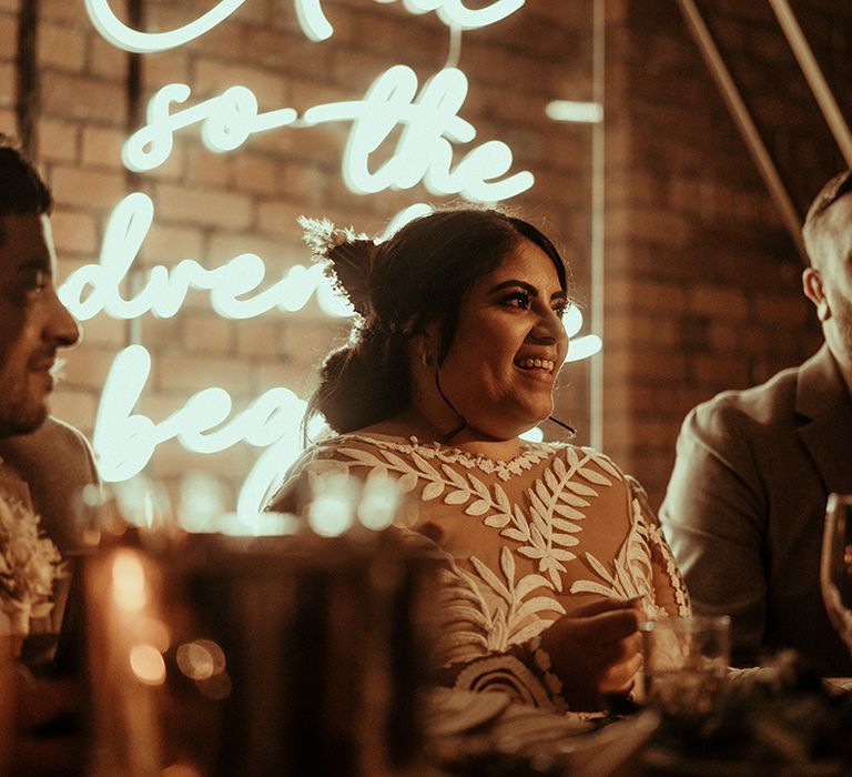 Bride in a lace wedding dress sitting at the top table with a neon sign behind the top table 