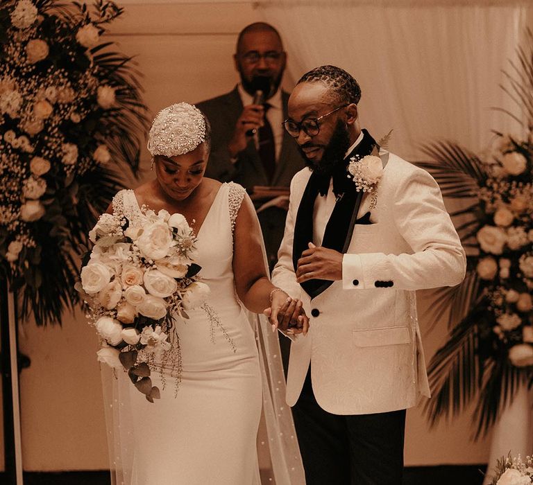 Black groom in white tuxedo jacket holding hands with his bride in a fitted Pronovias wedding dress and beaded headdress as they walk down the aisle as husband and wife 