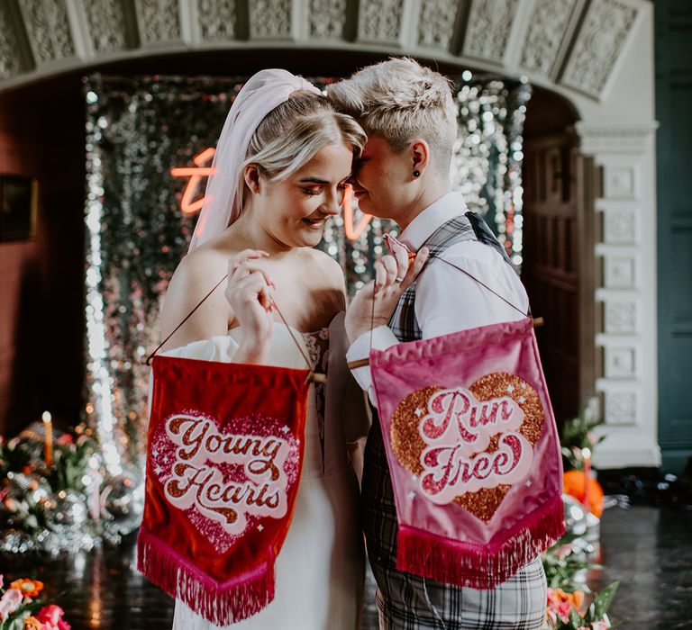 Two brides holding velvet fabric banners saying young hearts run free at fun retro wedding 