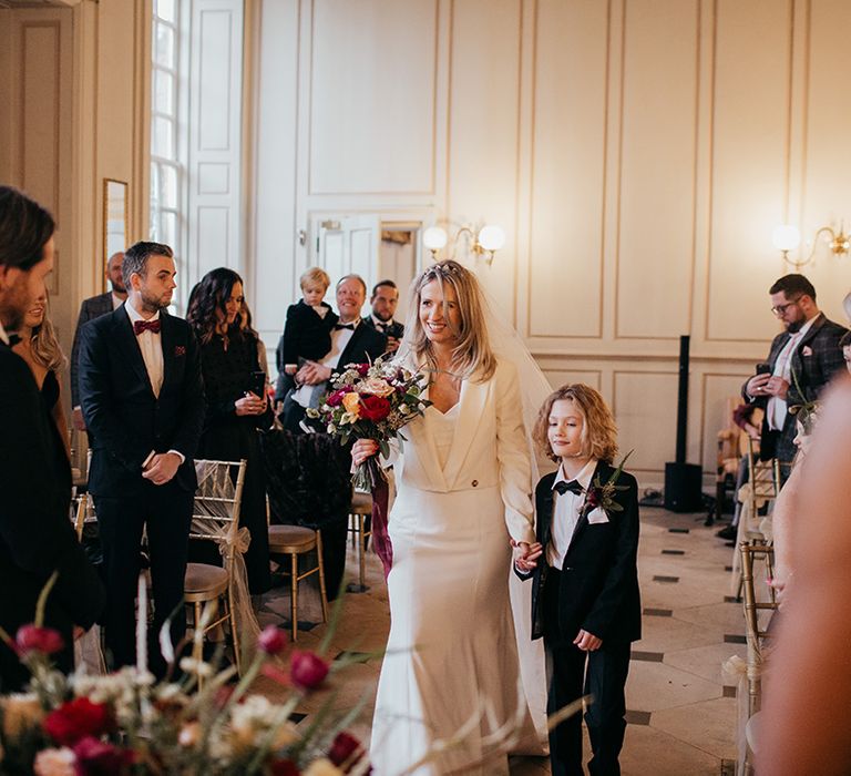 Bride walks down the aisle with son who wears black tie as bride wears Zara jacket and Suzanne Neville wedding gown 