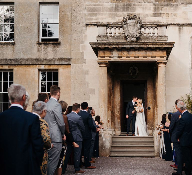 Bride and groom share a kiss in doorway of Elmore Court wedding venue in front of wedding guests