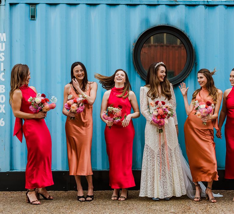 Bride in lace buttoned rixo dress with bridesmaids in red and orange satin dresses in various styles