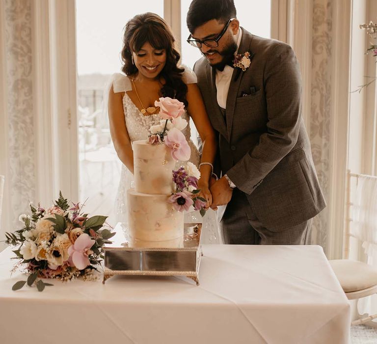 Bride and groom cutting into two tier neutral coloured wedding cake with mixed pinks and floral decorations