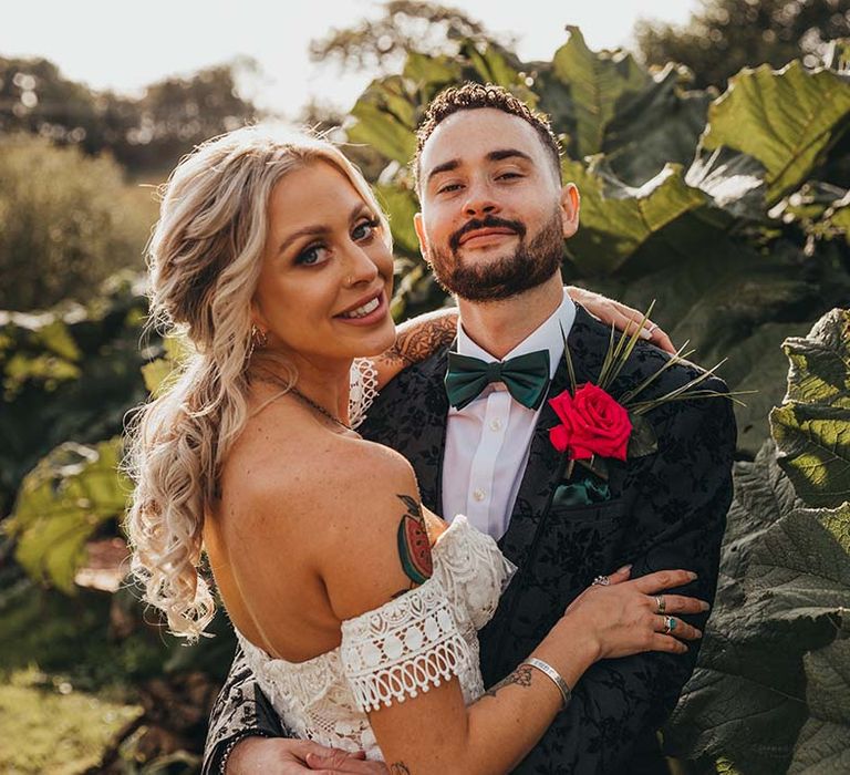 Groom in dapper black three piece patterned suit with red rose boutonnière and dark green bow tie with bride in strapless lace boho wedding dress 