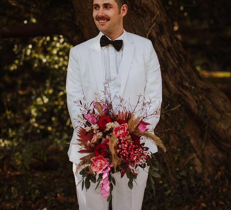 Groom in a three-piece white groom suit with black bow tie holding a Brian red and pink wedding bouquet 